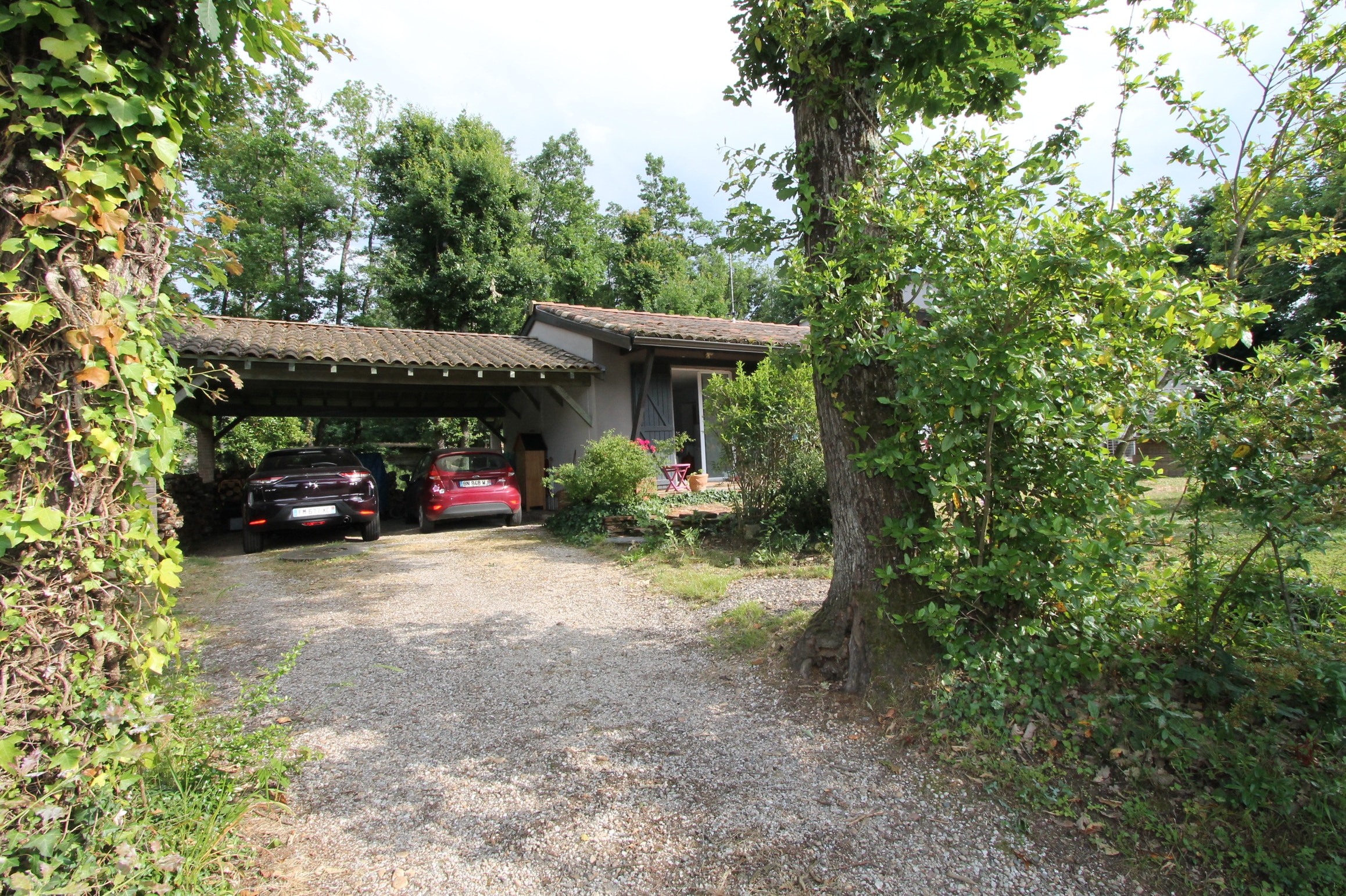 Allée de gravier menant à un gîte de campagne avec abri voiture tuilé. Deux voitures sont garées près de l'entrée entourée de verdure et d'arbres.