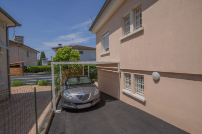 Abri voiture moderne adossé à une maison rose pâle. Une petite voiture grise est garée sur l'allée bitumée sous un ciel bleu d'été.