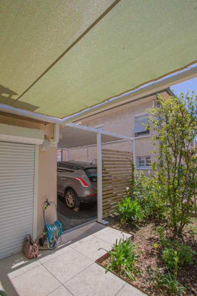 Voile d'ombrage vert couvrant une terrasse en dalles et un abri voiture moderne avec claustra bois. Jardin résidentiel ensoleillé.