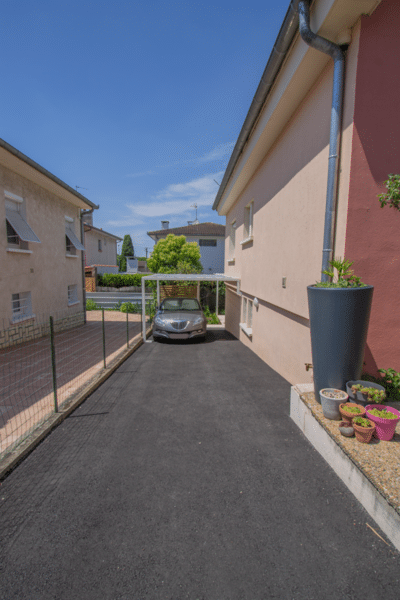 Allée d'accès pavillonnaire asphaltée encadrée par deux maisons. Un carport moderne abrite une berline grise sous un ciel bleu.