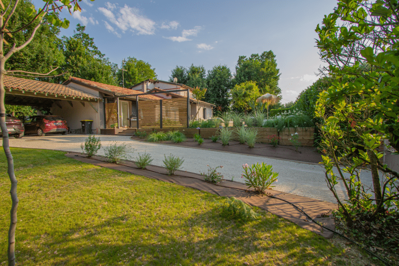Maison avec carport et pergola en bois, visible depuis une longue allée en gravier. Pelouse verte et jardin paysager surélevé de graminées ornementales.