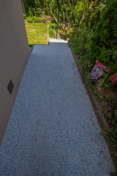 Long passage latéral entre un mur beige et une haie, recouvert de gravier résine gris-bleu clair. Le chemin mène à un jardin verdoyant.