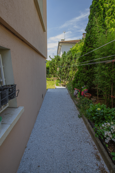Allée latérale résidentielle en gravier blanc, bordée par une façade saumon et de hautes haies. Pots sur l'appui de fenêtre.