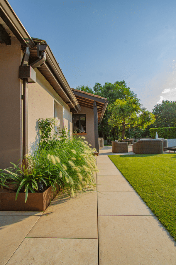 Allée pavée le long d'une maison beige avec une jardinière en acier Corten. Graminées ornementales luxuriantes face à la pelouse verte.