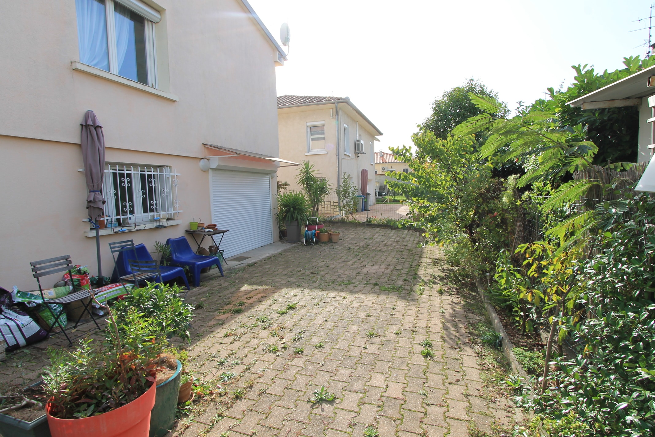 Allée pavée d'une maison de banlieue, envahie de mauvaises herbes. Végétation luxuriante, chaises bleues et garage visible sur le mur beige.