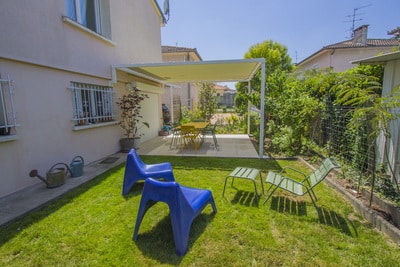 Jardin ensoleillé, pelouse verte, chaises bleues et salon de jardin. Pergola moderne abritant une terrasse avec table jaune, près de la maison.