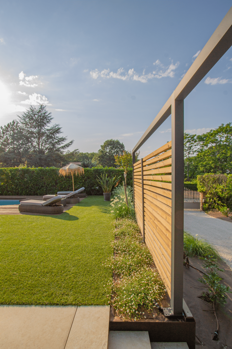 Jardin moderne avec clôture en lattes de bois et structure métallique. Pelouse verte, transats, massif floral et allée de gravier.