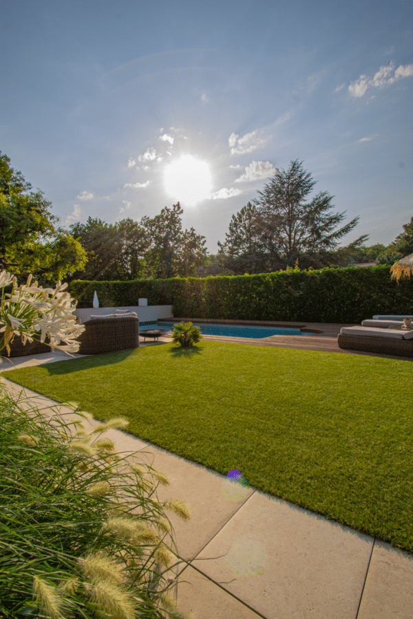 Aménagement de luxe pour jardin avec piscine entourée d'une terrasse en bois. Gazon vert, mobilier moderne et soleil éclatant.