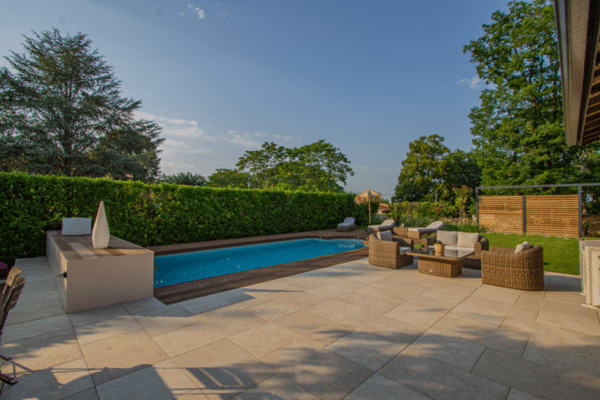 Piscine moderne et longue terrasse carrelée, aménagée avec un salon de jardin en rotin tressé. Haie végétale dense en arrière-plan.