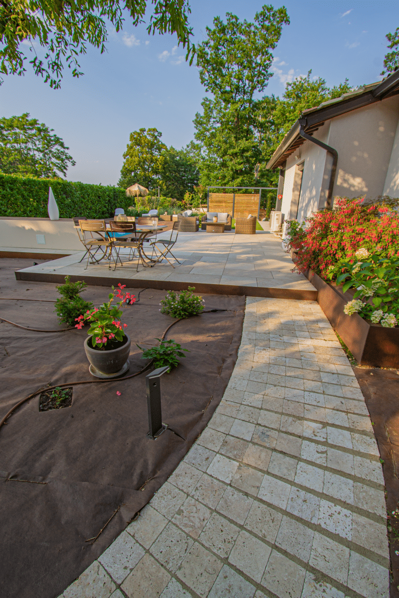Chemin pavé vers une terrasse moderne avec mobilier extérieur lounge. Jardin en aménagement bordé de jardinières Corten fleuries.