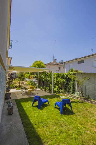 Vue d'un jardin ensoleillé avec pelouse, menant à une terrasse ombragée par une pergola blanche. Table extérieure jaune et chaises longues bleues.