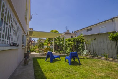 Jardin résidentiel ensoleillé avec pelouse, deux transats bleus et une terrasse couverte par une pergola. Espace repas extérieur et verdure.