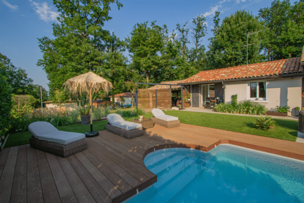 Aménagement extérieur avec piscine, grande terrasse en bois et transats. Maison de vacances en arrière-plan sous un ciel bleu et des arbres.