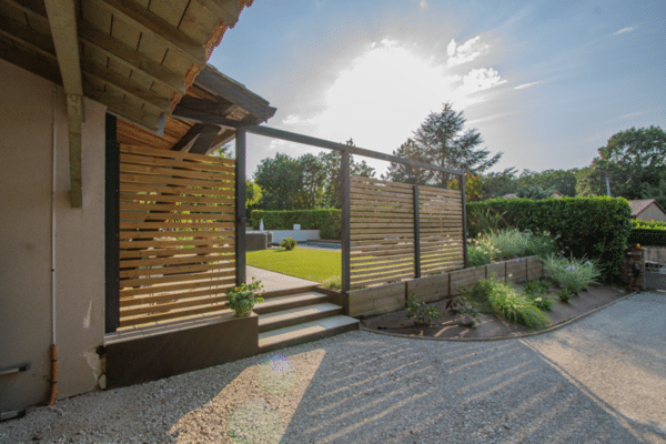 Aménagement extérieur : claustras ajourés en bois, escalier en béton et massif paysager, séparant l'allée en gravier du jardin et de la piscine.
