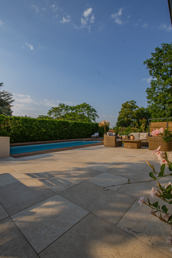 Grande terrasse en dalles de travertin clair avec piscine, haie verte, et salon de jardin en rotin sous un ciel d'été.