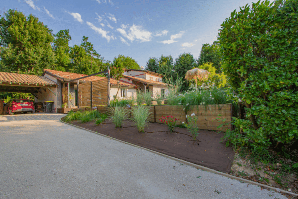 Façade de villa avec carport, voiture rouge et allée en gravier. Jardin paysager frais avec jardinières en bois, graminées et parasol en paille.