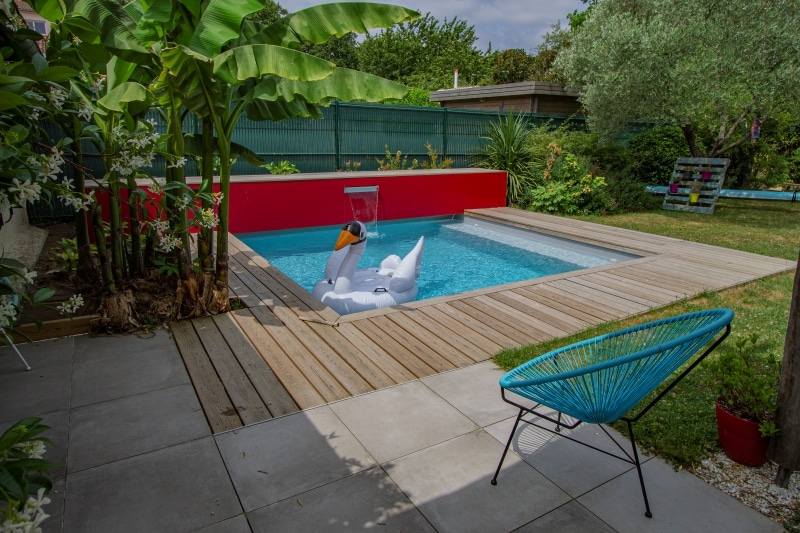 Mini-piscine moderne, terrasse en bois, mur rouge et cascade. Jardin tropical avec bananiers et fauteuil Acapulco turquoise.