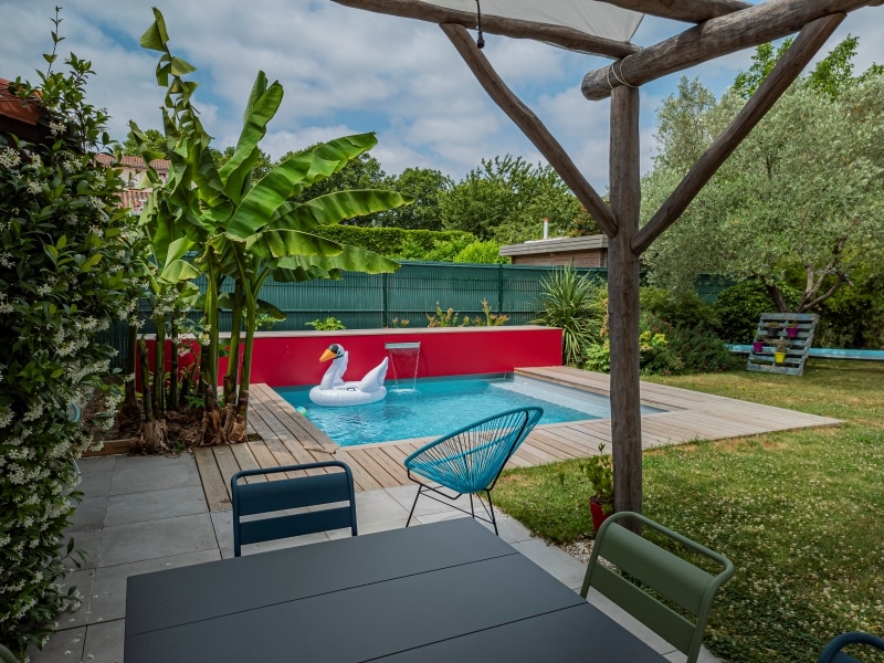 Terrasse ombragée près d'une mini-piscine moderne. Mobilier extérieur design, grand bananier, mur rouge vif et cygne gonflable.