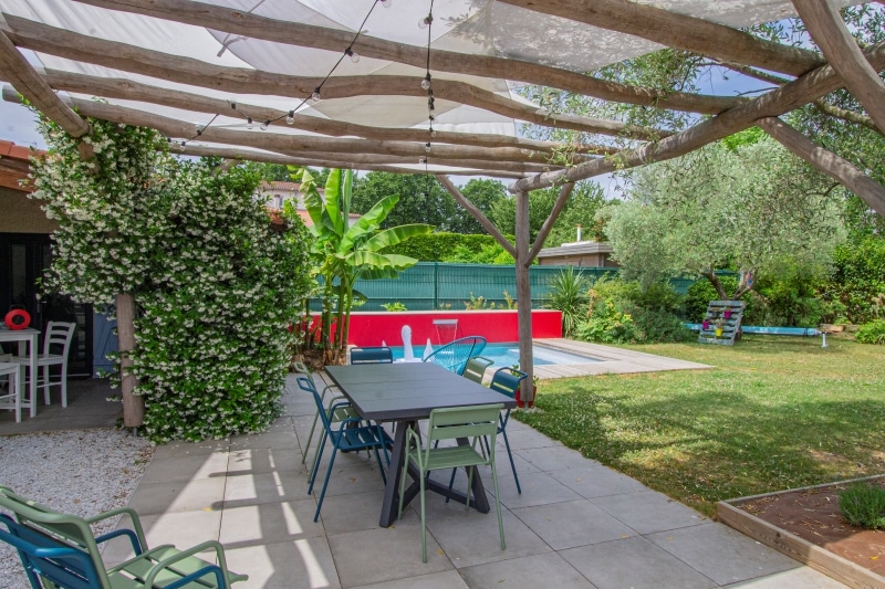 Terrasse sous pergola en bois brut avec table et chaises design colorées. Vue sur le jardin, la piscine et le jasmin en fleurs.