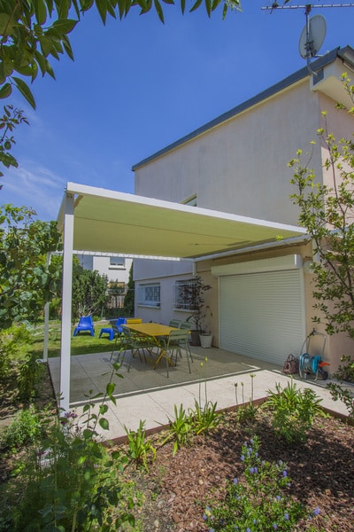 Pergola blanche couvrant une terrasse pavée. Table jaune et mobilier extérieur, entourée d'un jardin luxuriant sous un ciel bleu d'été.