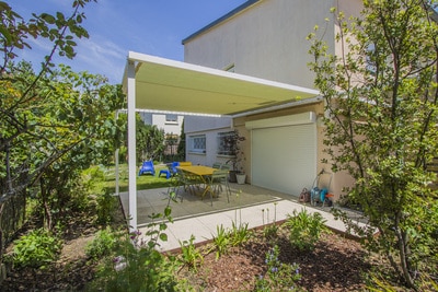 Aménagement extérieur avec une pergola créant de l'ombre sur la terrasse pavée. Table à manger et jardin luxuriant sous un ciel bleu.