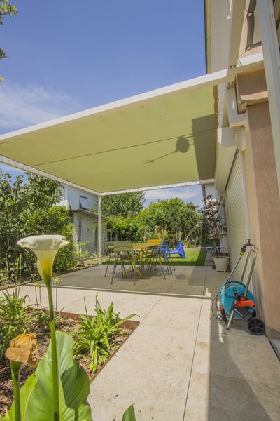 Pergola moderne avec toile tendue jaune sur une terrasse ensoleillée. Mobilier extérieur coloré, carrelage clair et lys blanc en premier plan.