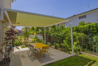 Terrasse carrelée et jardin ensoleillé sous une pergola à toile jaune. Table à manger extérieure jaune pour six, entourée de verdure.
