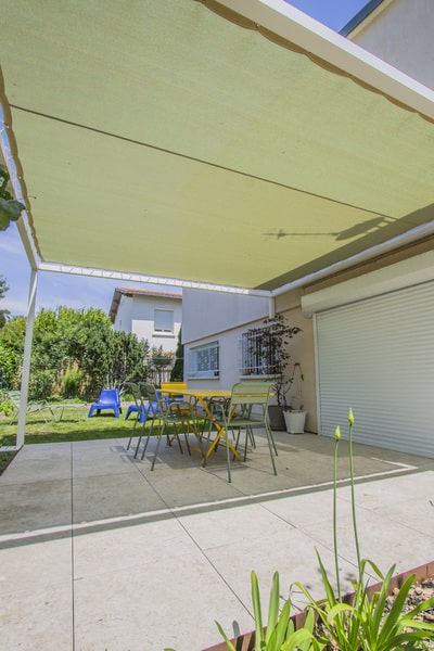 Terrasse moderne en dalles claires sous une pergola. Voile d'ombrage vert protégeant le salon de jardin jaune et vert du soleil.