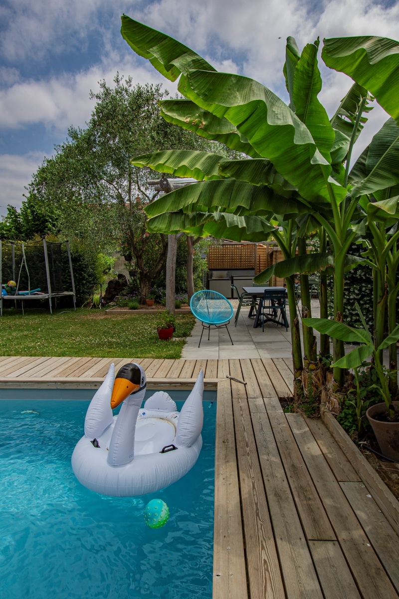 Piscine turquoise avec cygne gonflable, terrasse en bois et bananiers luxuriants, menant à un coin repas et un trampoline.