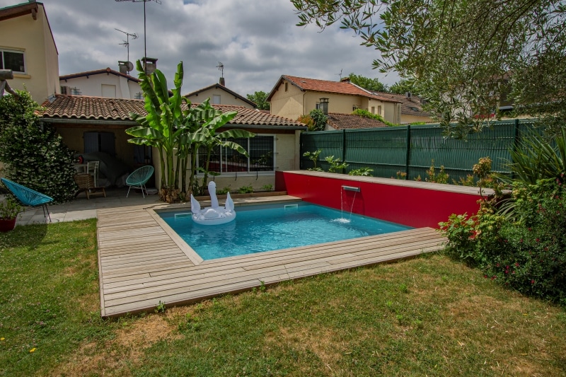 Piscine moderne avec terrasse en bois, mur rouge vif et cascade. Cygne gonflable, bananiers et patio aménagé dans ce jardin.