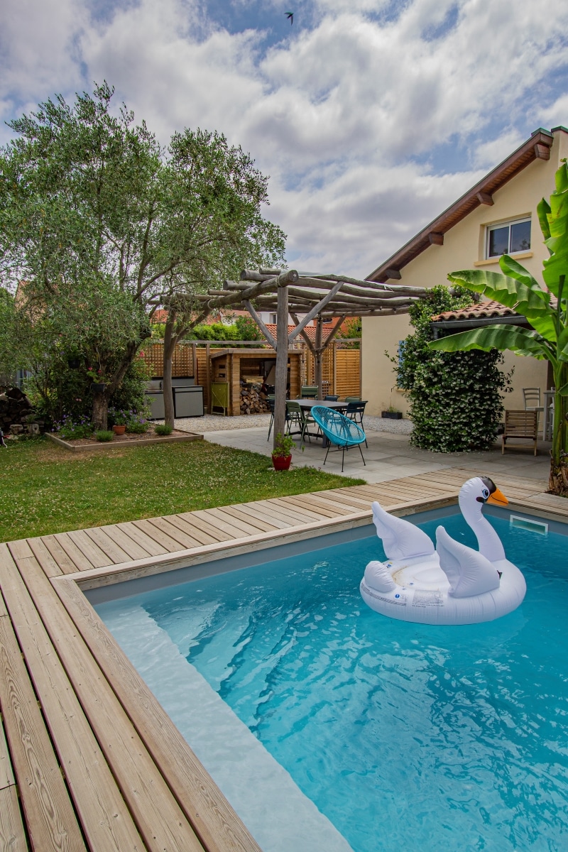 Piscine de jardin avec margelle en bois, cygne gonflable blanc, pergola rustique et coin repas sous un olivier.