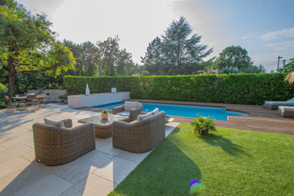 Aménagement extérieur luxueux: piscine, terrasse en bois, patio clair. Salon de jardin en rotin face à la pelouse et à la haie.