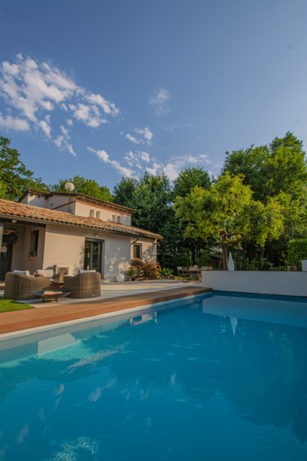 Vue plongeante sur une piscine bleue éclatante. Terrasse en bois et salon de jardin luxueux devant la villa. Entourée d'arbres sous un ciel d'été.
