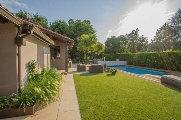 Magnifique aménagement extérieur avec longue piscine bordée de bois, terrasse moderne, mobilier en rotin et gazon vert devant une maison.