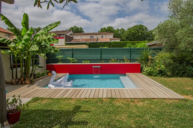 Piscine moderne avec deck bois, mur rouge vif, cascade minimaliste, bananiers et cygne gonflable dans un jardin ensoleillé.