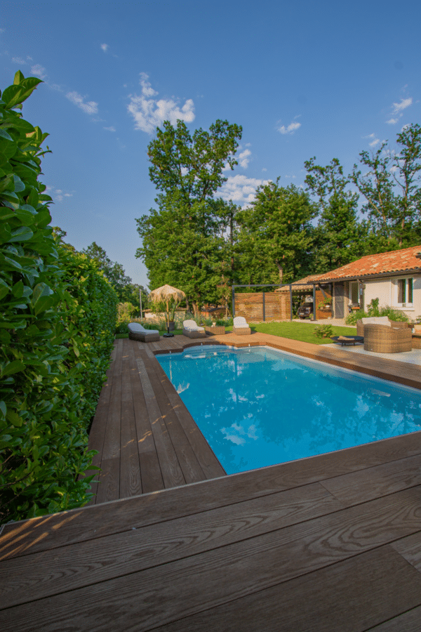 Piscine rectangulaire moderne, terrasse bois et haie verte. Vue plongeante sur le bassin, chaises longues et pool house sous un ciel bleu d'été.