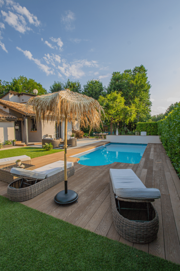 Piscine moderne et sa large terrasse en bois composite entourée d'un jardin luxuriant. Chaises longues et parasol paillote.