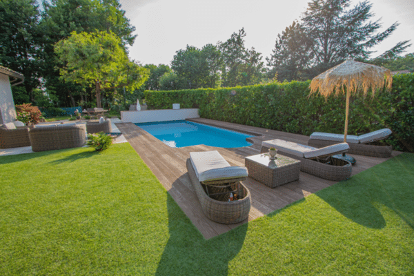 Jardin avec piscine sur terrasse bois, entourée de gazon luxuriant et d'une haie dense. Salon de jardin et transats pour la détente.