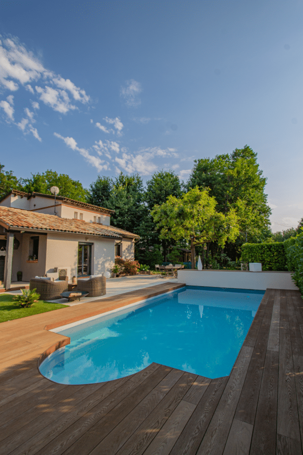 Piscine moderne et rectangulaire avec terrasse en bois composite cernant une maison style provençal sous un ciel bleu d'été. Salon de jardin.
