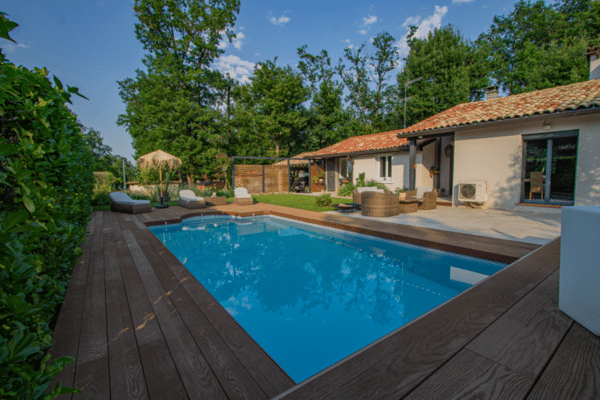 Piscine extérieure moderne, longue terrasse en bois composite sombre, mobilier de détente et maison de vacances sous un ciel bleu dégagé.
