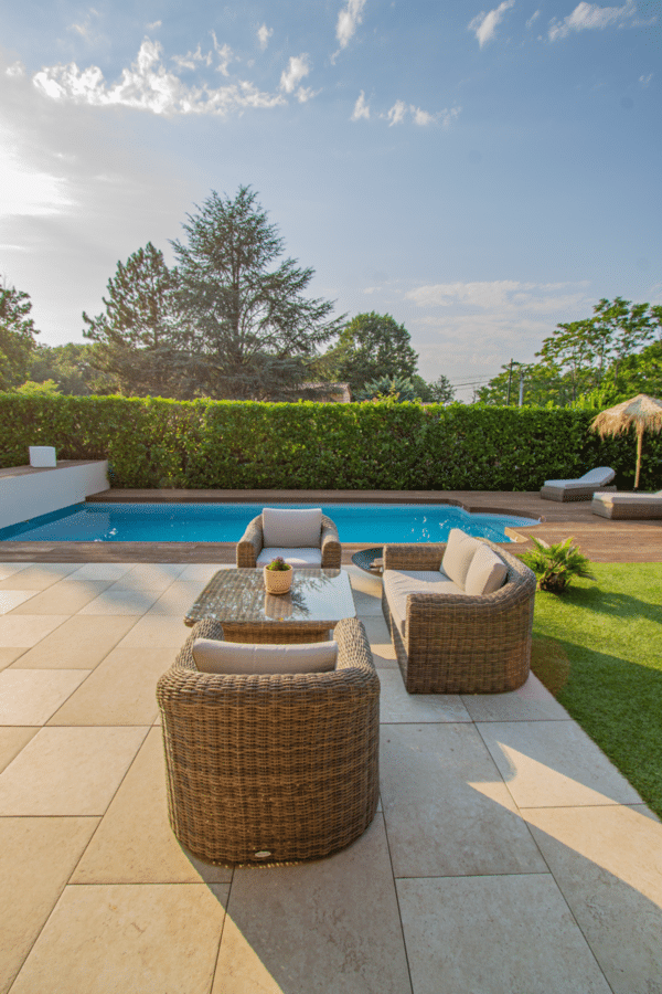 Terrasse luxueuse près de la piscine au coucher du soleil. Salon de jardin en osier sur grand carrelage de pierre beige, avec haie de verdure.