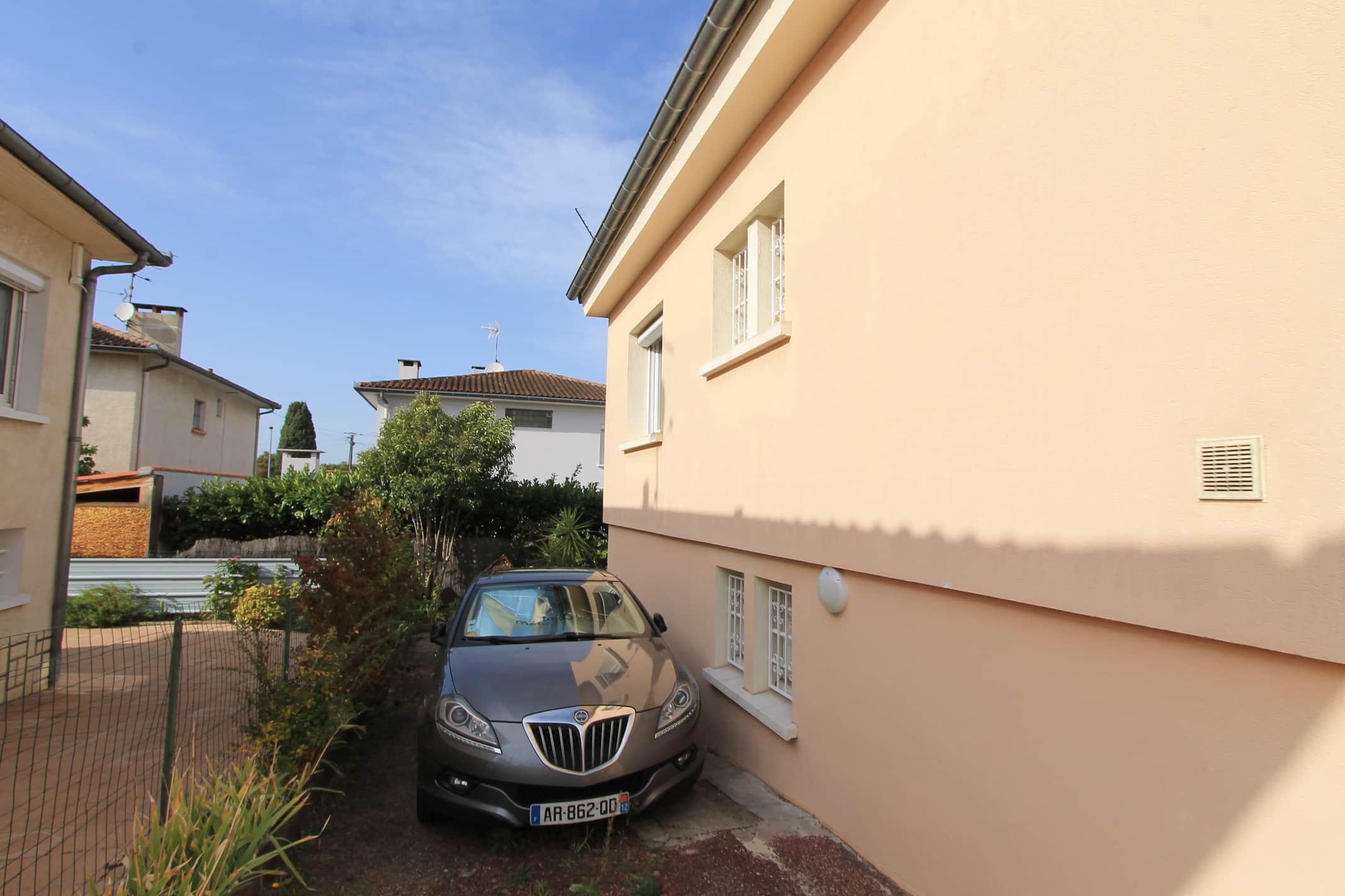 Façade couleur pêche d'une maison avec deux fenêtres à barreaux. Une Lancia Delta grise est garée en parking très étroit. Plaque AR-862-QD 12.