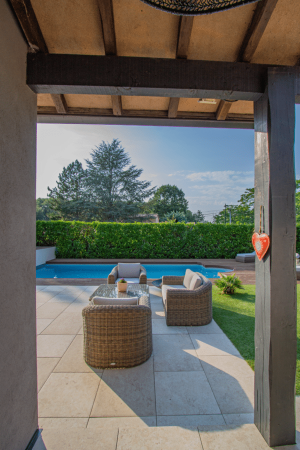 Salon de jardin en rotin sur une terrasse carrelée sous une pergola, avec vue sur la piscine et la haie luxuriante.