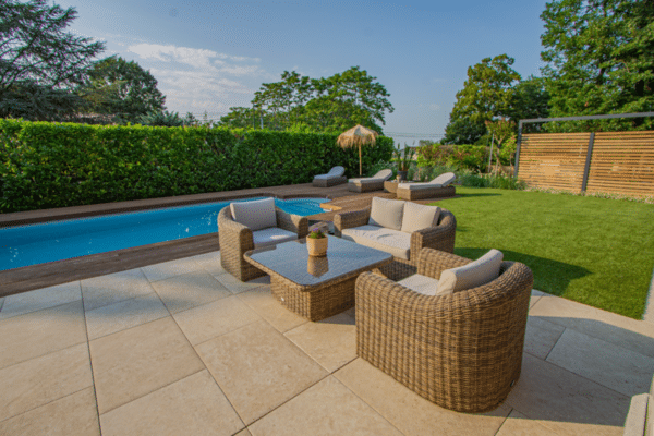 Terrasse en dalles beiges avec salon en rotin, bordant une piscine bleue et un deck en bois. Jardin paysager avec chaises longues.