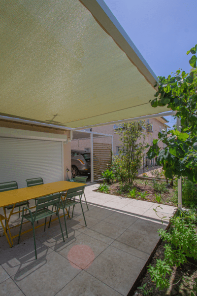 Patio ombragé sous pergola: coin repas (table jaune, chaises vertes) et jardin paysager. À l'arrière, un carport abrite une voiture.