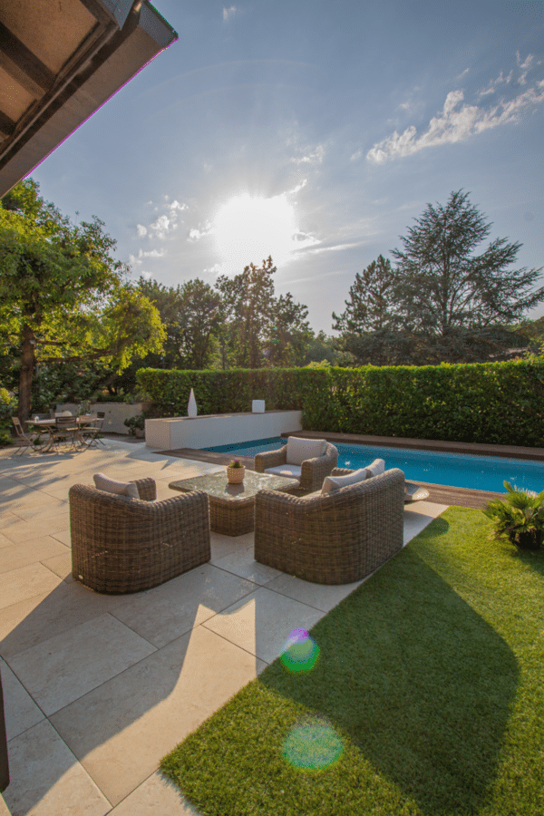 Grande terrasse en pierre avec salon de jardin en rotin près d'une piscine bordée de haies, éclairée par le soleil couchant.