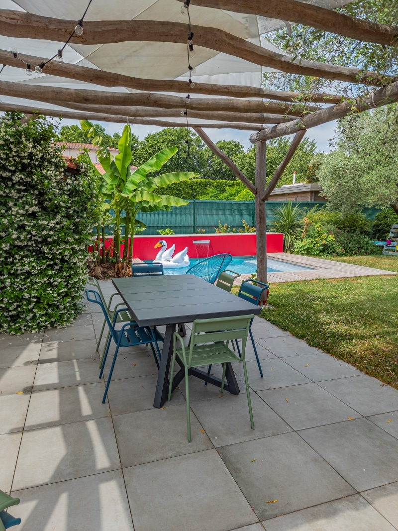 Salle à manger extérieure sous pergola en bois. Table grise et chaises colorées près d'une piscine avec cygne gonflable, mur rouge et bananier.