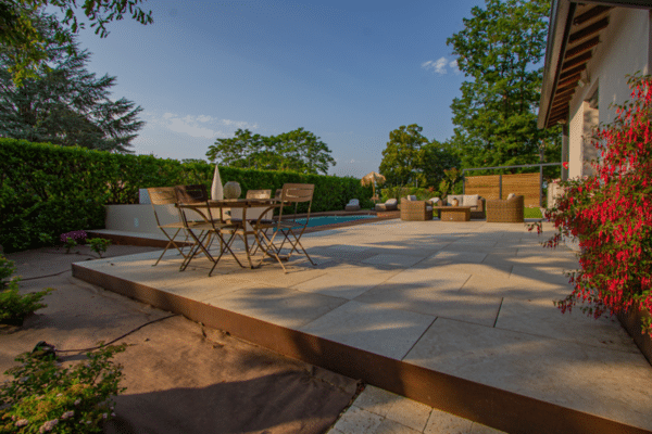 Terrasse moderne en pierre claire, bordure Corten. Salon de jardin, coin repas, et piscine. Ambiance extérieure luxueuse et verdoyante.
