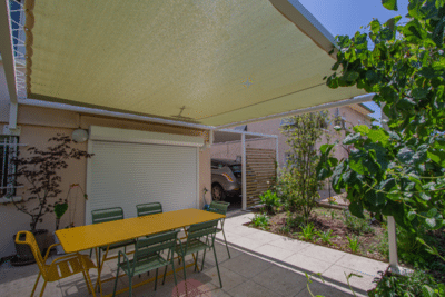 Terrasse de repas ombragée par un auvent, avec une table jaune et des chaises vertes. Vue sur le jardin verdoyant et le garage/carport.