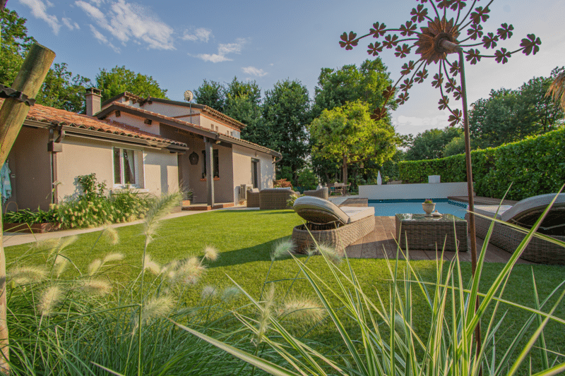 Maison et piscine de villa entourées d'un jardin luxuriant. Transats en rotin sur la terrasse en bois et graminées décoratives au premier plan.